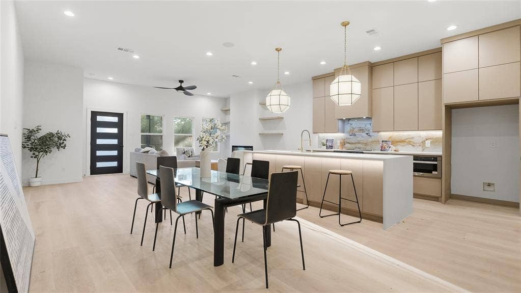 Open-concept interior featuring light wood-finish flooring, custom flat-panel cabinetry, a stone-finished backsplash, and a central island with under-cabinet lighting