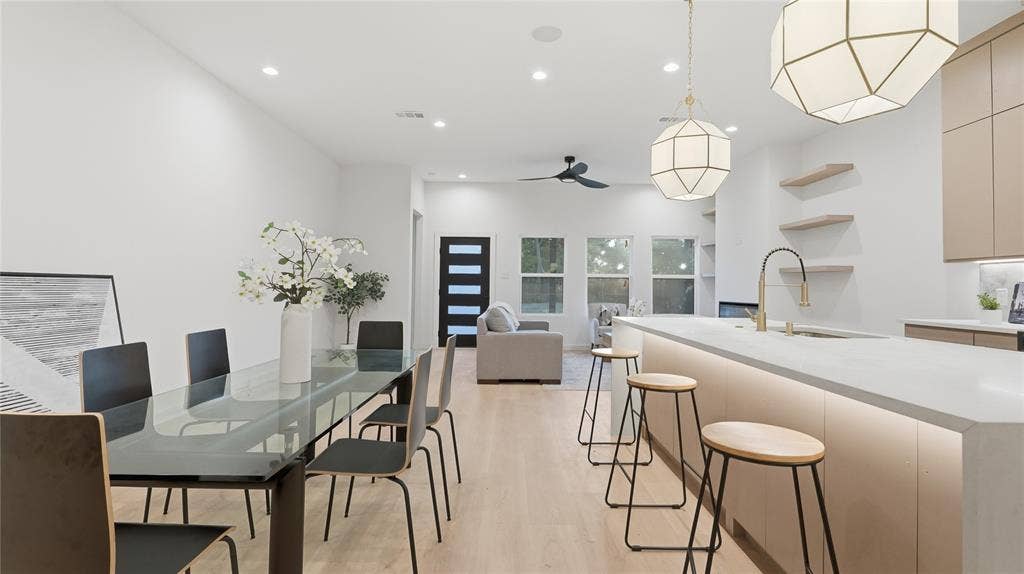 Open-concept space featuring light wood-finish flooring, a waterfall-edge island with integrated under-cabinet lighting, geometric pendant light fixtures, and minimalist built-in wall shelving