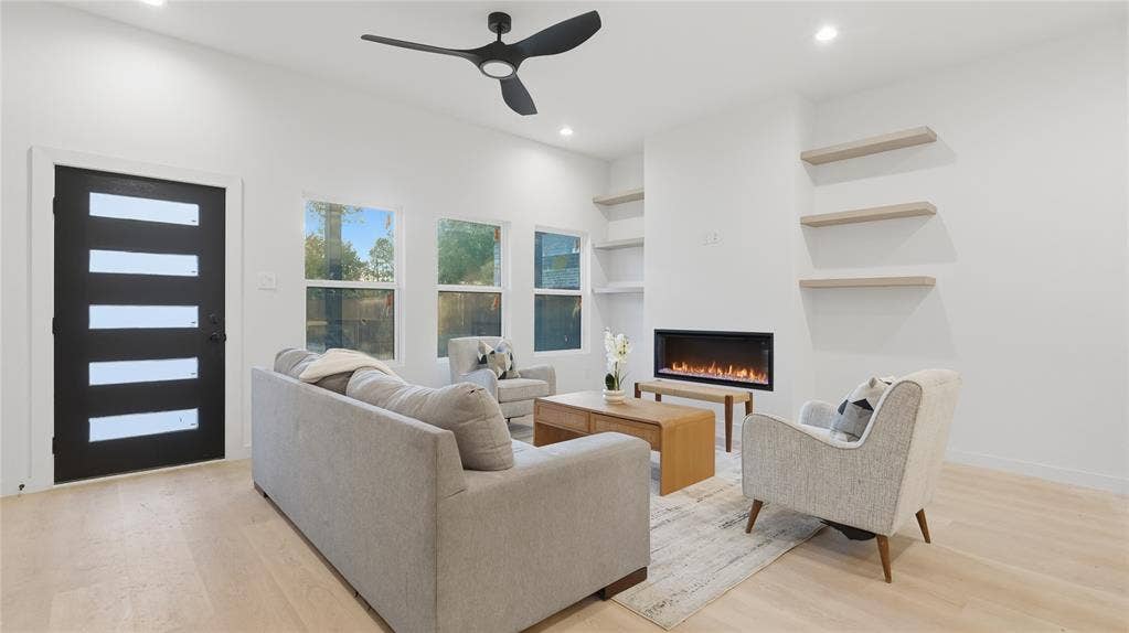 Modern interior featuring light-toned wood-finish flooring, a linear electric fireplace, recessed lighting, matte black ceiling fan, and built-in floating shelves