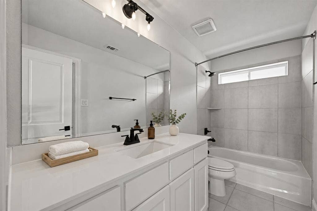 White vanity cabinetry with a stone countertop, integrated sink, large wall-mounted mirror, matte black plumbing fixtures, and a tub-shower combination featuring tiled wall surround