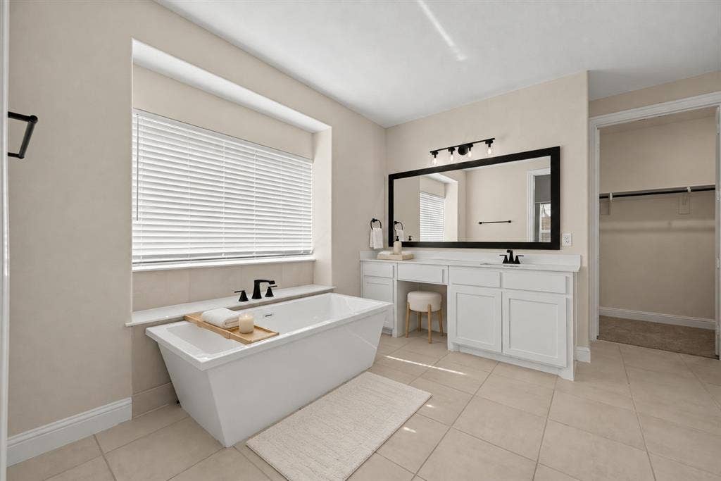 Freestanding soaking tub with matte black deck-mounted faucet, white cabinetry with integrated vanity seating, large rectangular mirror with black frame, tile flooring, and light-filtering horizontal blinds