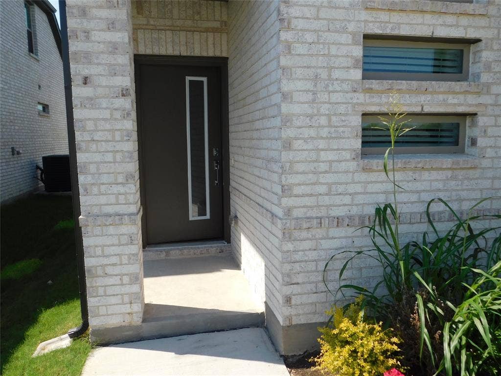 2702 Baynes Place, Princeton, TX 75407 Light-toned brick exterior featuring a recessed entry, modern dark entry door with a vertical glass insert, and stacked rectangular windows