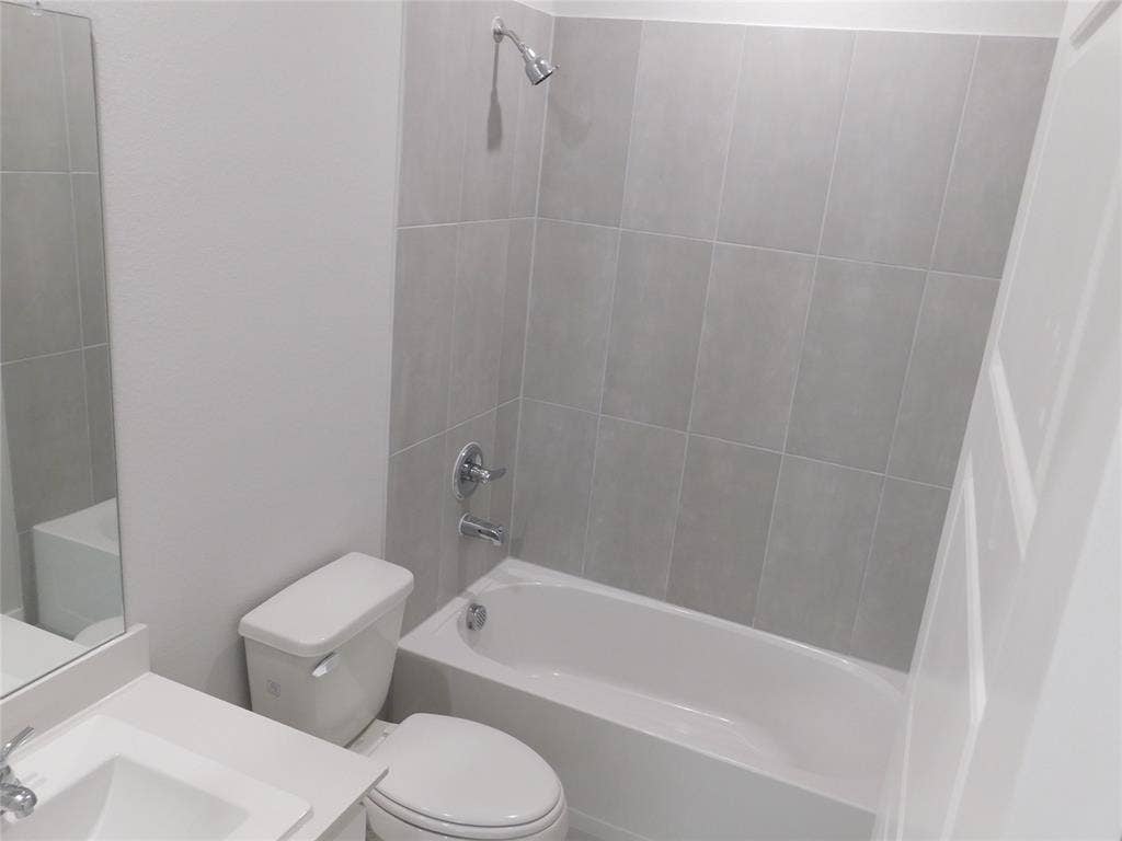 2702 Baynes Place, Princeton, TX 75407 Bathroom featuring a porcelain tub and shower combination with full-height grey tile surround, white solid-surface vanity countertop, undermount sink with chrome hardware, and wall-mounted vanity mirror
