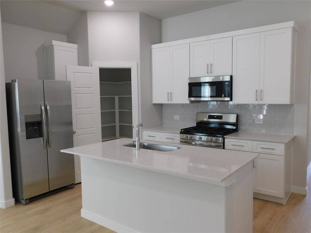 2702 Baynes Place, Princeton, TX 75407 Kitchen featuring white shaker cabinetry, quartz countertops, a central island with an integrated stainless steel sink, stainless steel appliances, and wood-finish flooring