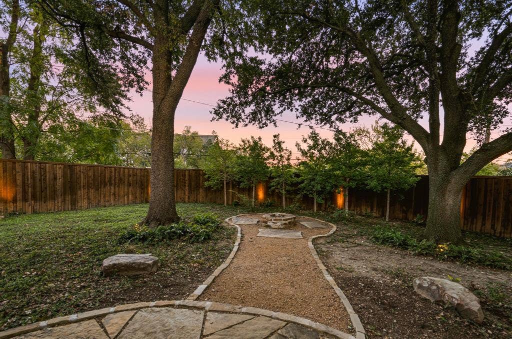 9656 Ravensway Drive, Dallas, TX 75238 Backyard featuring a stone-lined gravel path, stone fire pit, wood privacy fencing, and mature trees