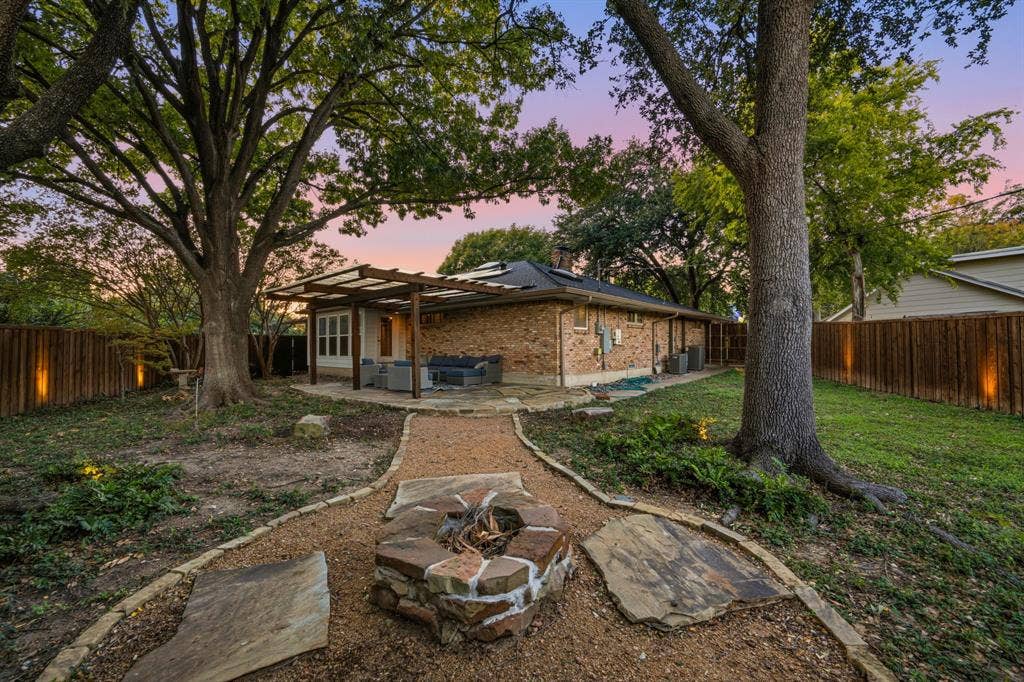 9656 Ravensway Drive, Dallas, TX 75238 Backyard featuring a stone-clad fire pit, a covered pergola, a natural stone path, mature shade trees, and perimeter wood fencing with integrated accent lighting
