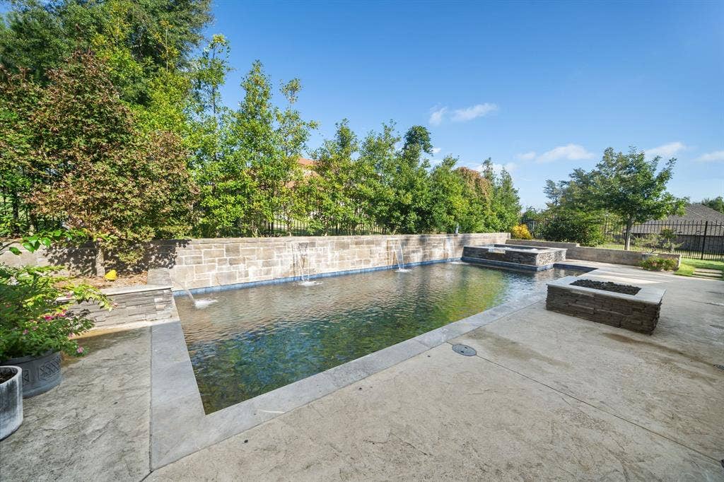7609 Timber Trail, Tyler, TX 75703 Rectangular swimming pool with integrated water features and a stone-clad retaining wall, accompanied by a raised spa, stone fire feature, and concrete patio