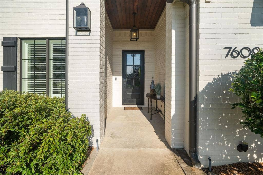 7609 Timber Trail, Tyler, TX 75703 Covered entryway featuring a painted brick facade, wood-plank ceiling, black metal-framed front door with glass inlay, and structured exterior lantern lighting