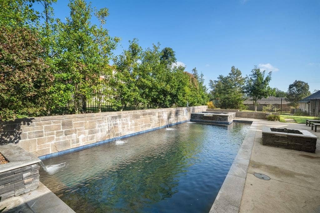 7609 Timber Trail, Tyler, TX 75703 Outdoor area featuring a rectangular pool with integrated water features, raised stone retaining wall, stone-surround spa, and a stone fire pit set on a concrete patio