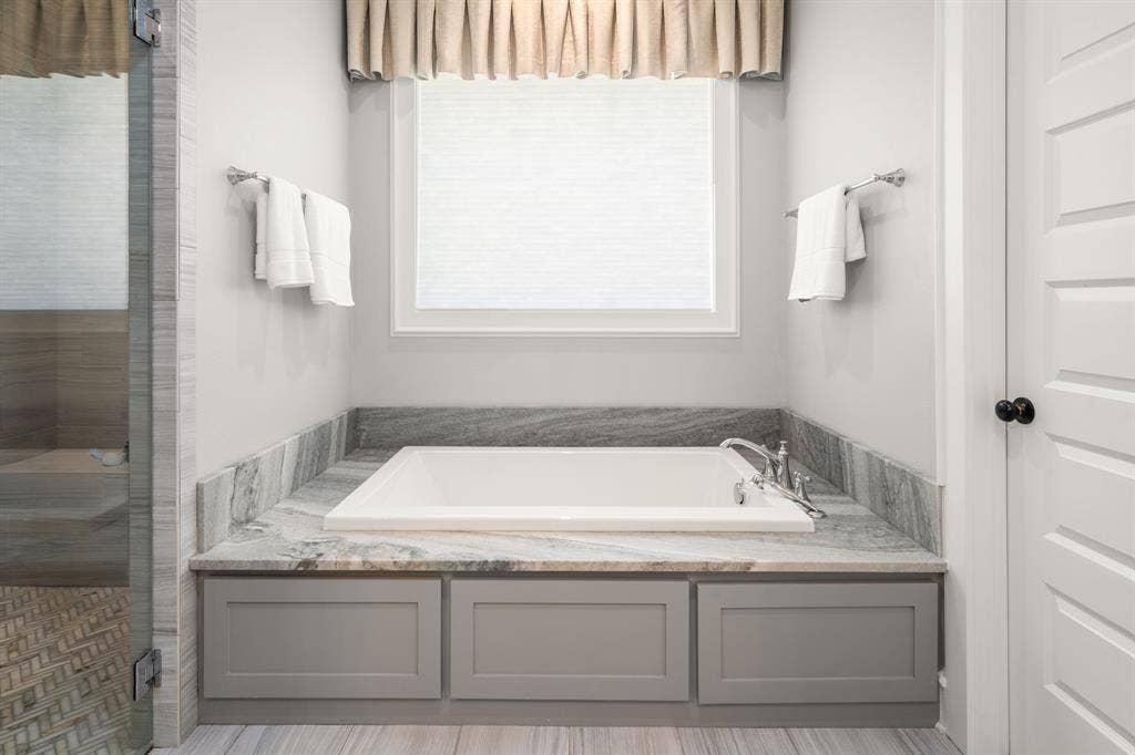 7609 Timber Trail, Tyler, TX 75703 Soaking tub with stone surround, custom gray cabinetry, chrome deck-mounted faucet, and window with neutral window treatment