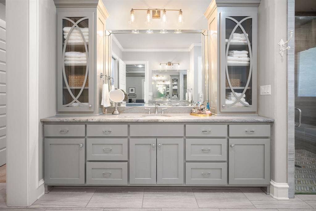 7609 Timber Trail, Tyler, TX 75703 Dual-sink vanity featuring gray shaker-style cabinetry, integrated glass-front shelving, a natural stone countertop, and an oversized polished-edge mirror with multi-bulb vanity lighting