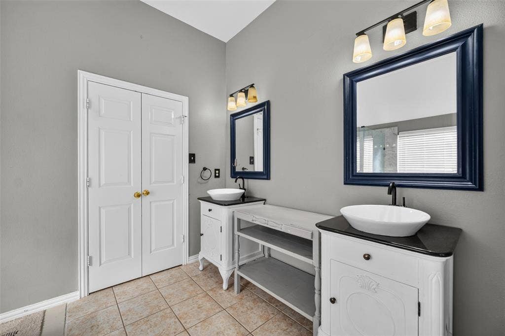 5726 Remington Drive, Garland, TX 75044 Dual vanities featuring white cabinetry, dark stone-finish countertops, ceramic vessel sinks, and dark metal faucets, accented by wall-mounted lighting and matching framed mirrors