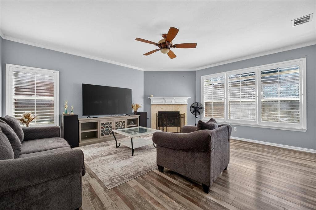 5726 Remington Drive, Garland, TX 75044 Living room featuring wood-finish flooring, a fireplace with a mantel, crown molding, a ceiling fan, and large windows with integrated blinds