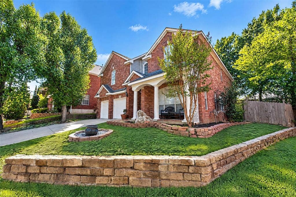 5726 Remington Drive, Garland, TX 75044 Traditional brick exterior featuring a covered front porch with white columns, two-car garage, and tiered natural stone retaining walls