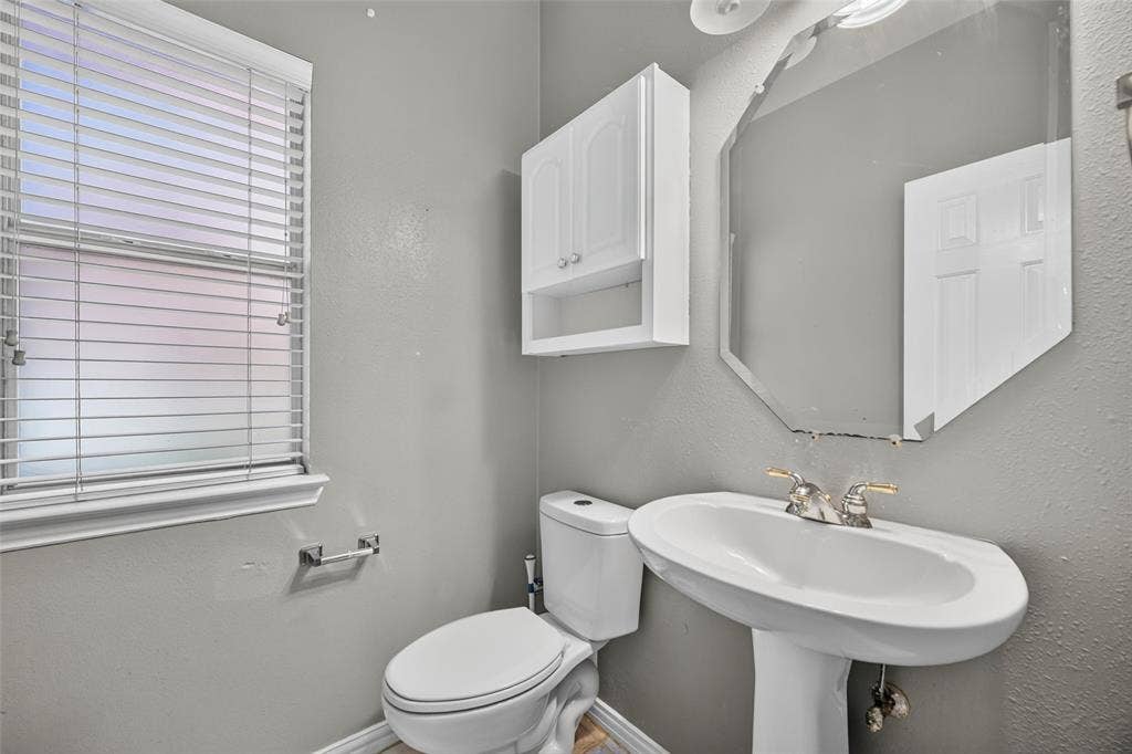 5726 Remington Drive, Garland, TX 75044 White pedestal sink with polished metal faucet, octagonal wall-mounted mirror, white wall-mounted cabinet, and horizontal window blinds