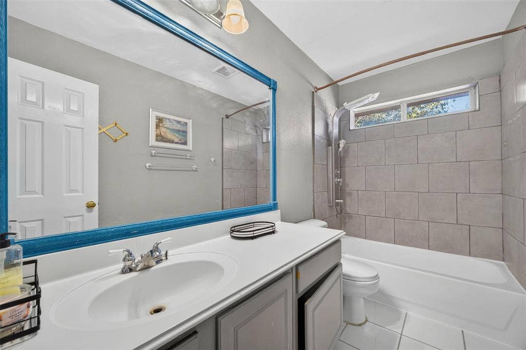 5726 Remington Drive, Garland, TX 75044 Bathroom featuring a white vanity with a solid-surface countertop, an inset basin, a full-width mirror, tile-surround shower-tub combination, and a transom window