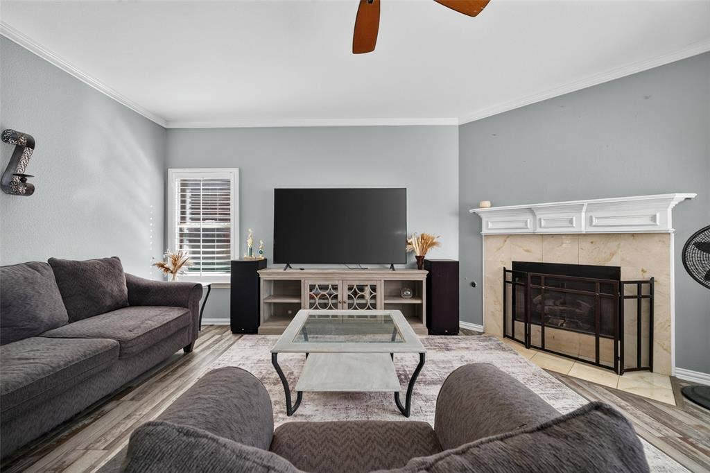 5726 Remington Drive, Garland, TX 75044 Living area featuring wood-finish flooring, a fireplace with a stone-surround and white mantel, crown molding, plantation shutters, and a ceiling fan
