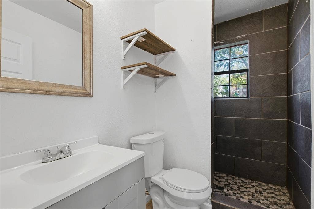 336 Bonnie View Road, Dallas, TX 75203 Bathroom featuring a vanity with a white countertop, wall-mounted floating wood-finish shelving, and a walk-in shower with dark large-format wall tiles and a mosaic-tile floor