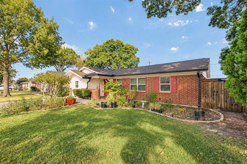 1910 R Avenue, Plano, TX 75074 Single-story brick exterior featuring a shingled gable roof, red shutters, and an expansive lawn with manicured landscaping and wood fencing
