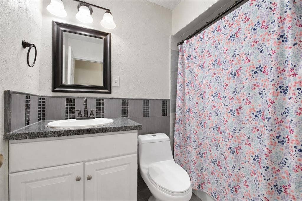 1910 R Avenue, Plano, TX 75074 White vanity cabinet with a stone-finish countertop, oval undermount sink, dark-toned mosaic tile backsplash, and a three-light vanity fixture