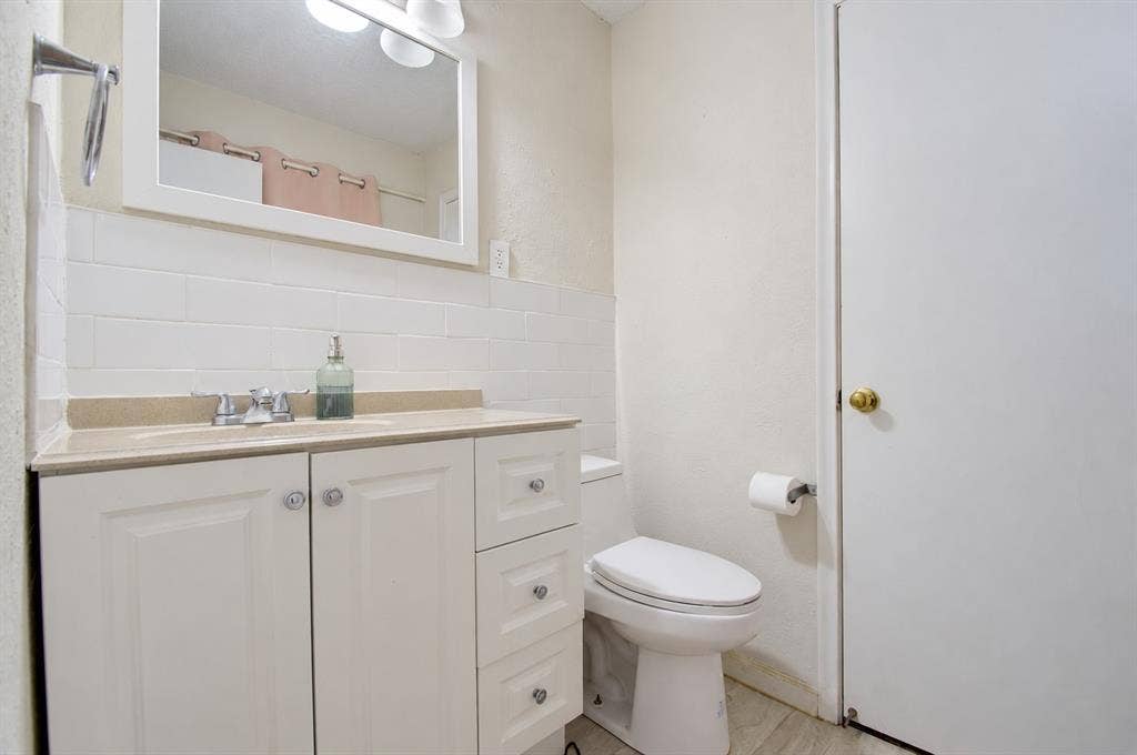 1910 R Avenue, Plano, TX 75074 White vanity cabinet featuring decorative hardware, rectangular countertop with integrated sink, white subway tile backsplash, wall-mounted mirror, and polished chrome fixtures