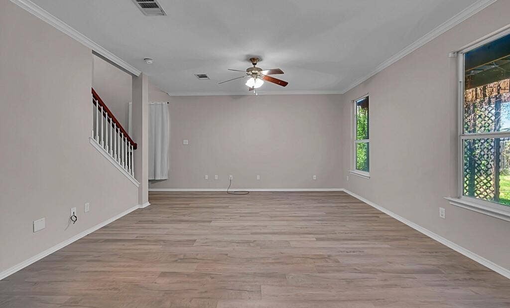 Neutral-toned interior featuring wood-finish flooring, crown molding, a ceiling fan with integrated lighting, and a staircase with a wooden handrail and white balusters