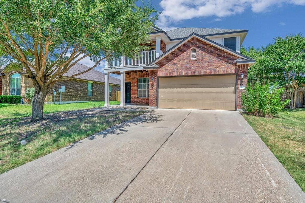 Two-story brick exterior featuring a covered front porch, a second-level balcony with metal railings, a concrete driveway, and an attached two-car garage