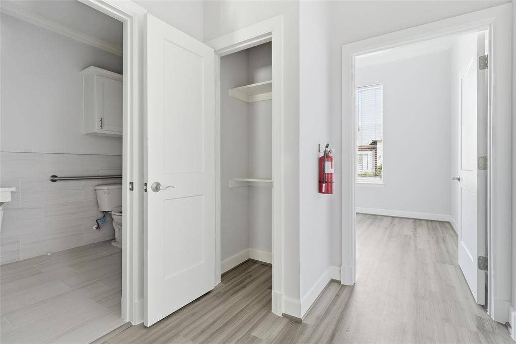 Interior hallway featuring wood-finish flooring, two-panel interior doors, recessed storage shelving, and light-toned tile flooring in the tiled room