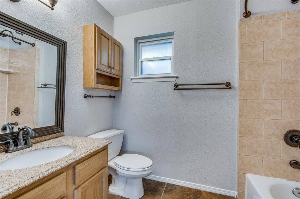 2131 Onetta Drive, Irving, TX 75061 Bathroom featuring a vanity with a stone-finish countertop and an integrated sink, framed wall mirror, wood-finish wall-mounted cabinetry, a tiled tub surround, and dark-toned metal hardware