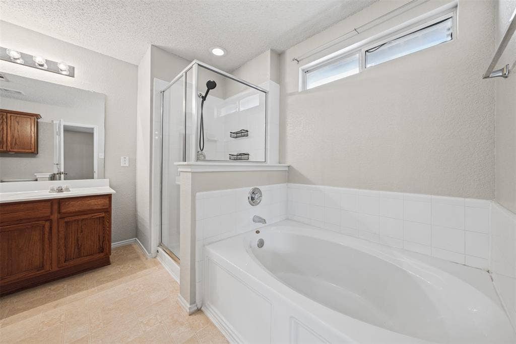 5428 Maverick Drive, Grand Prairie, TX 75052 Bathroom featuring a wood-toned vanity with a white countertop, oval soaking tub with white tile surround, separate glass-enclosed shower, and clerestory window