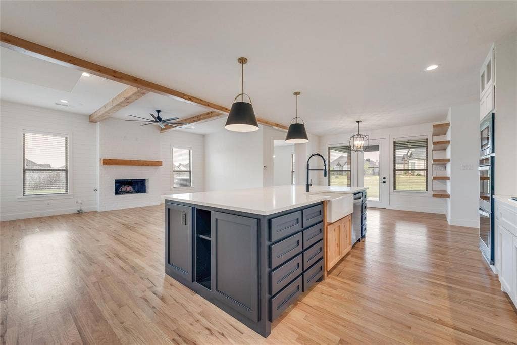 1001 Devoss Court, Caddo Mills, TX 75135 Open-concept kitchen featuring an island with stone-finish countertops, apron-front sink, and custom cabinetry, complemented by wood-finish flooring, exposed ceiling beams, and a fireplace with a wood mantel