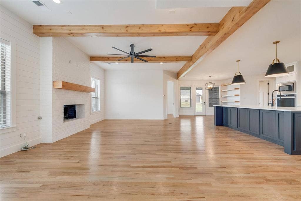 1001 Devoss Court, Caddo Mills, TX 75135 Open-concept interior featuring exposed wooden ceiling beams, wood-finish flooring, a white brick fireplace with a wood mantel, and kitchen island with built-in cabinetry and pendant lighting