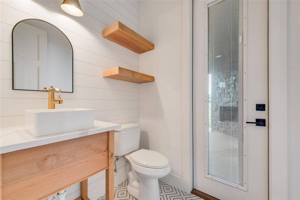 1001 Devoss Court, Caddo Mills, TX 75135 Custom wood-finish vanity with a white rectangular vessel sink, arched mirror, decorative shiplap wall detail, and geometric-patterned tile flooring