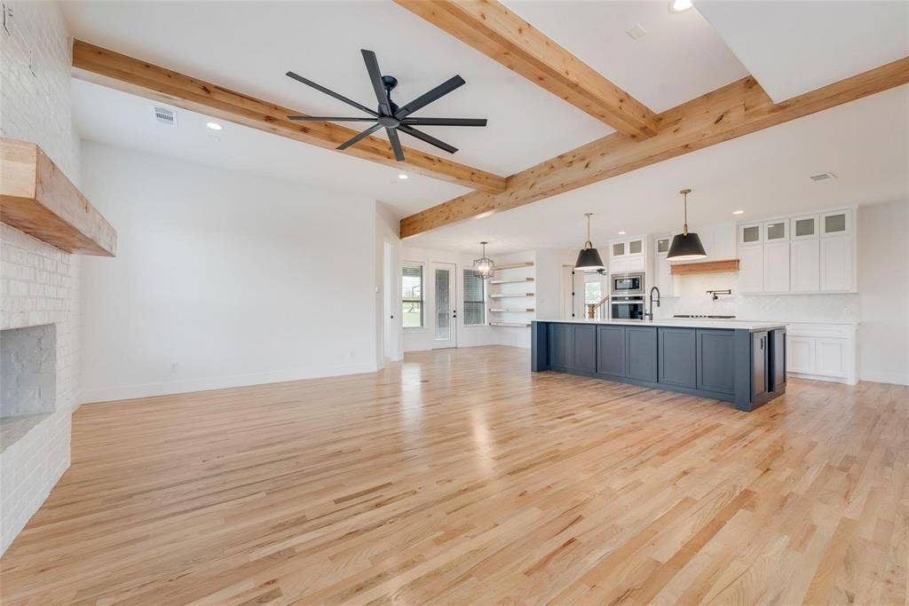 1001 Devoss Court, Caddo Mills, TX 75135 Open-concept interior featuring wood-finish flooring, exposed wooden ceiling beams, a painted brick fireplace with a wooden mantel, a large kitchen island with contrasting cabinetry, and floating wall shelves