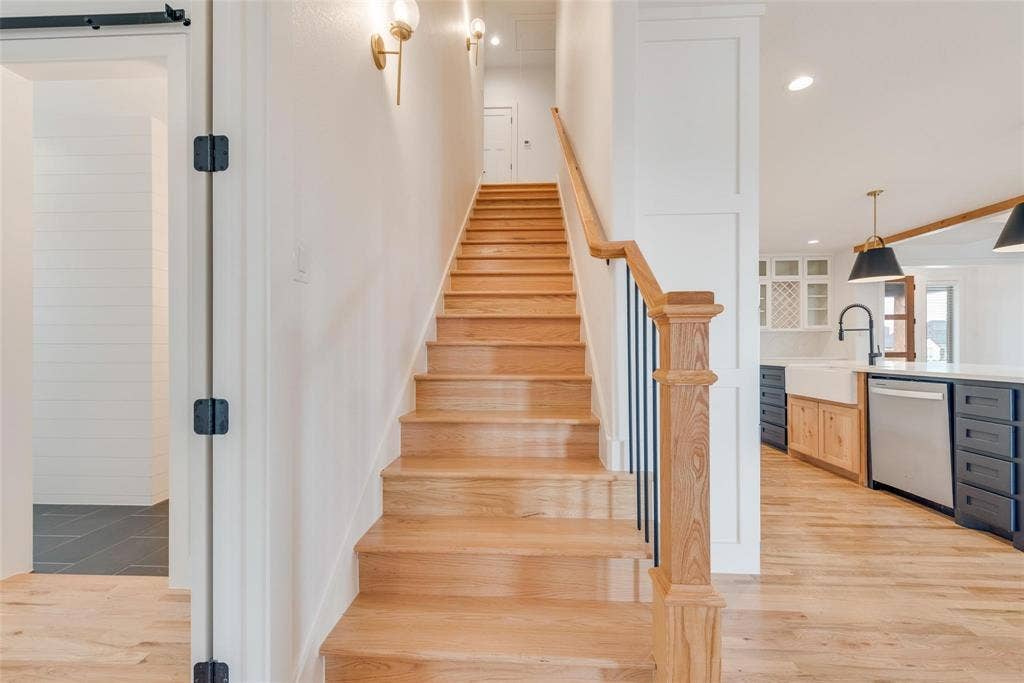 1001 Devoss Court, Caddo Mills, TX 75135 Staircase featuring wood-finish treads and a handrail with black metal balusters, complemented by wall-mounted sconce lighting, wood-finish flooring, and shaker-style cabinetry with a farmhouse-style sink
