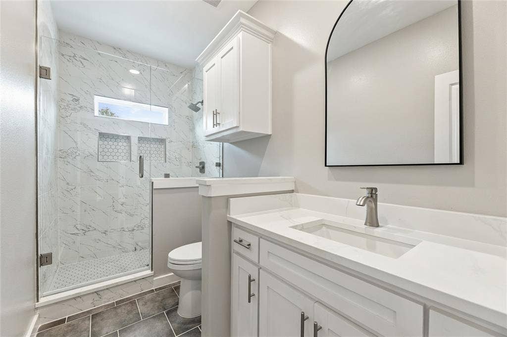 1242 Seminole Drive, Dallas, TX 75217 Walk-in shower with stone-finish tile surround and built-in niches, white cabinetry with countertop-integrated sink, arched wall mirror, and grey tile flooring