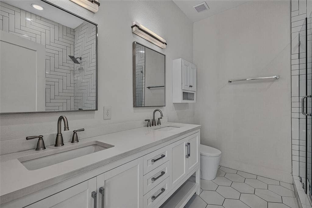 2222 Moser Avenue, #105, Dallas, TX 75206 Dual-sink vanity with quartz countertop, white shaker-style cabinetry, matte black hardware, glass-enclosed shower with herringbone tile detailing, and hexagonal floor tiling