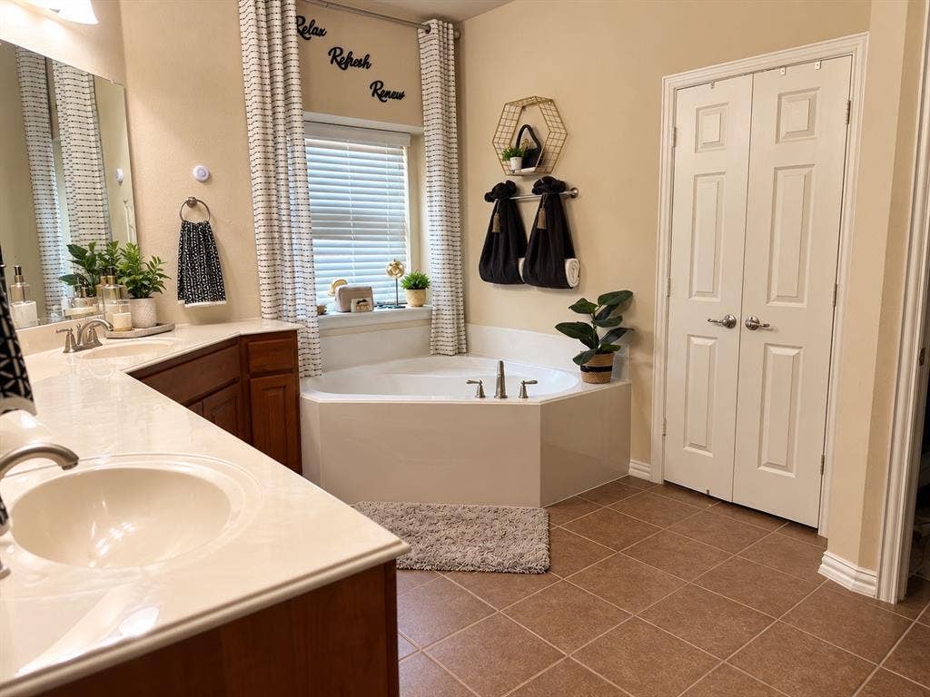 Bathroom featuring a double-sink vanity with light-toned countertops, a corner soaking tub with chrome fixtures, and tile flooring