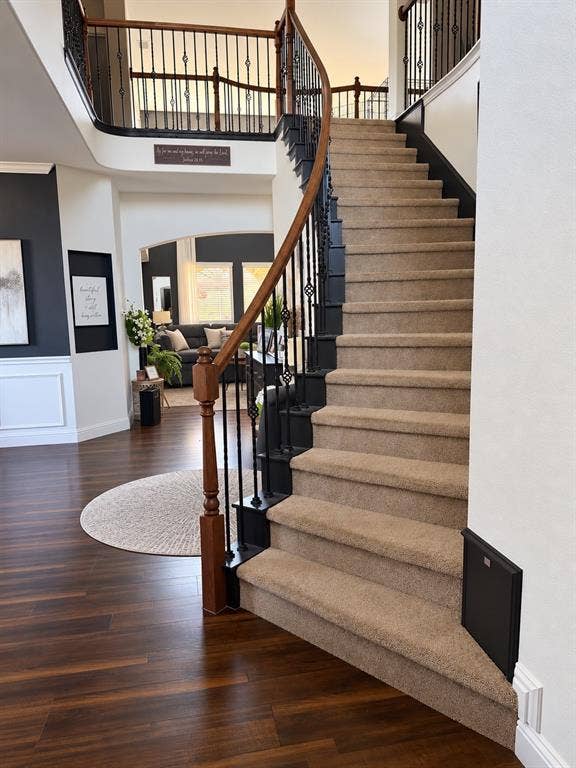 Curved staircase featuring carpeted treads, wood-finish handrails, and metal balusters, complemented by wood-finish flooring and wall wainscoting