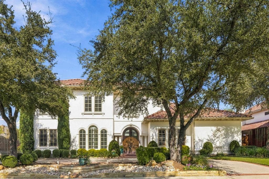 Mediterranean-inspired exterior featuring stucco facade, terracotta tile roof, arched entryway with decorative wood doors, multi-pane arched windows, and mature shade trees