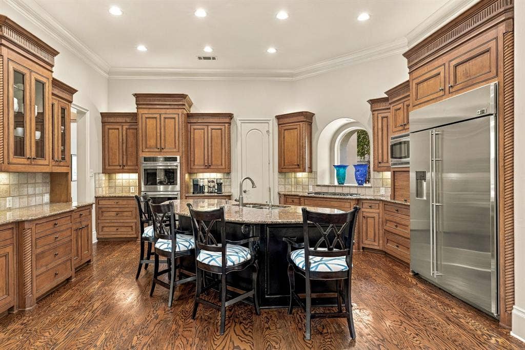 Kitchen featuring custom wood cabinetry, central island with an integrated sink, stainless steel appliances, stone countertops, and wood-finish flooring