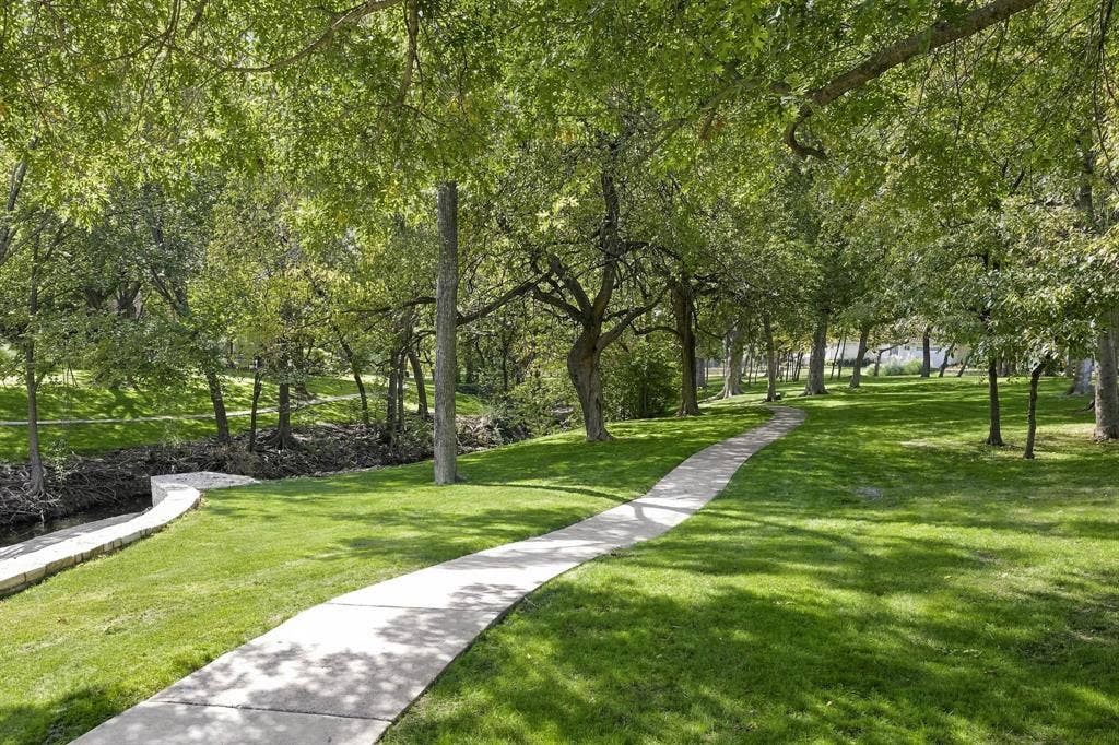 Expansive grounds featuring mature shade trees, a winding concrete pathway, stone retaining wall, and a natural creek border