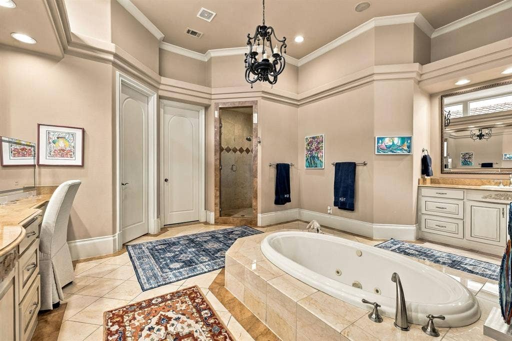 Built-in vanity cabinetry with stone countertops, a central jetted tub with stone tile surround, ornate chandelier lighting, and a walk-in shower with tile surround