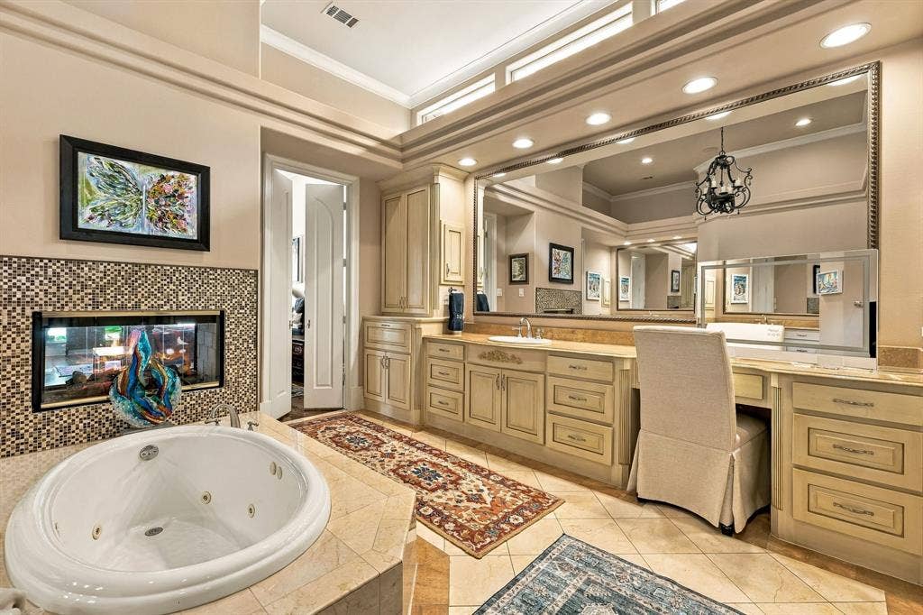 Jetted soaking tub with tile surround, double-sided fireplace with mosaic tile detailing, dual-sink vanity with custom cabinetry, expansive wall-mounted mirror, and recessed lighting