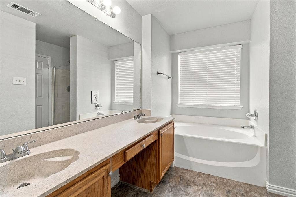 Dual-sink vanity with solid surface countertop, wood cabinetry, full-width wall mirror, soaking tub, and stone-finish flooring