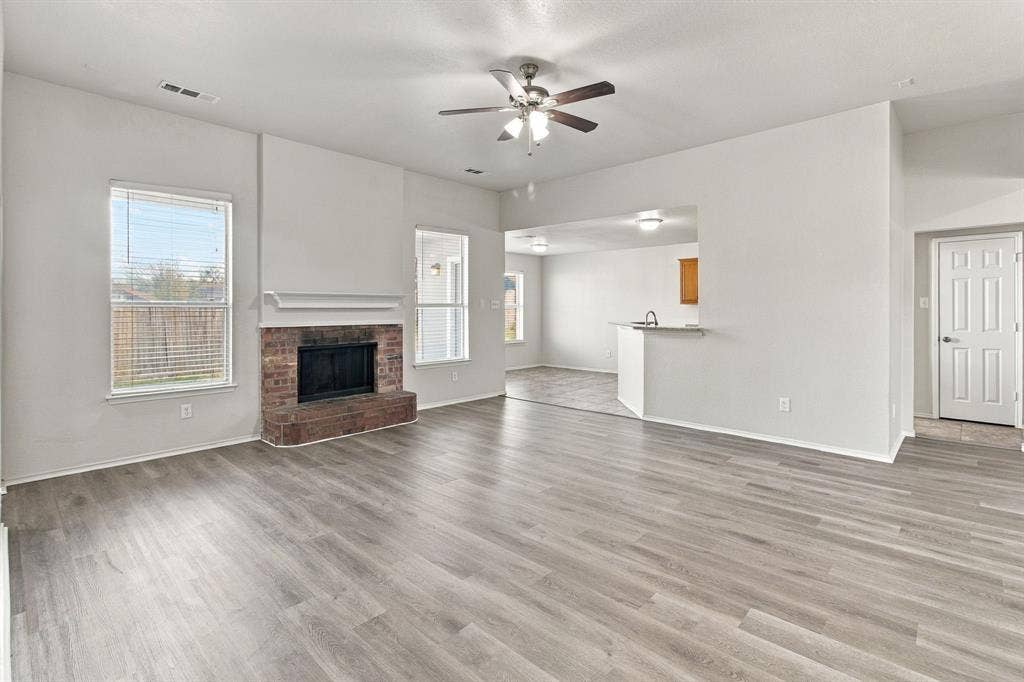 Open-concept living space featuring wood-finish flooring, a brick fireplace with a mantle, dual-aspect windows, and a ceiling fan with integrated lighting