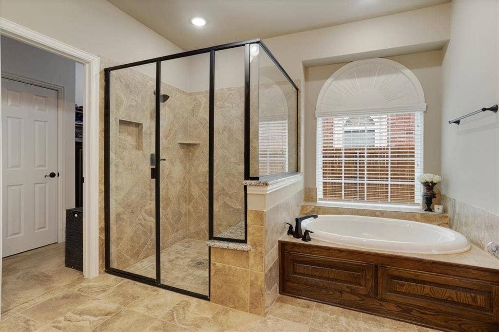 319 Westphalian Drive, Celina, TX 75009 Bathroom featuring a frameless glass shower enclosure with dark bronze accents, a built-in soaking tub with dark wood paneling, and a large arched window with blinds