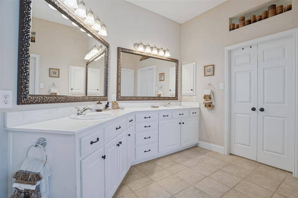 701 Crabapple Way, McKinney, TX 75072 Dual-sink vanity with white cabinetry, light-toned countertop, and decorative framed mirrors, complemented by ceramic tile flooring and recessed wall shelving