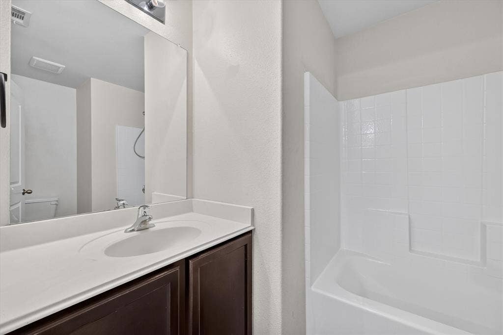 14276 Greenhaw Lane, Dallas, TX 75253 Bathroom featuring a dark wood-finish vanity, integrated white countertop with a single-basin sink, polished chrome faucet, full-width mirror, and a white tiled bathtub surround