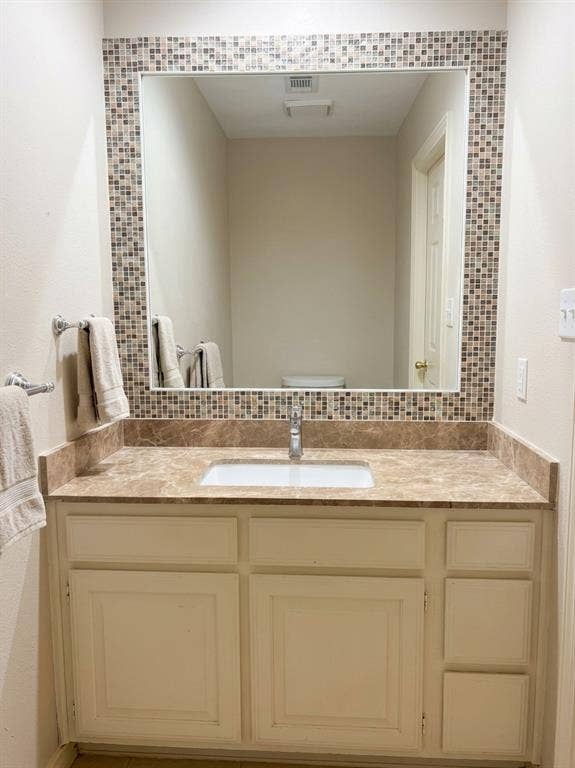 6011 Highplace Circle, Dallas, TX 75254 Bathroom vanity featuring white cabinetry, a rectangular undermount sink, stone-finish countertop with matching backsplash, and a large mirror framed by decorative mosaic tile